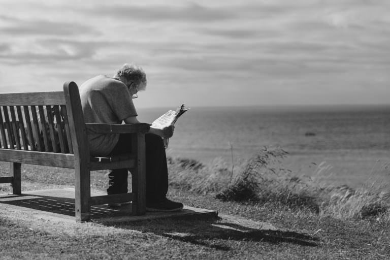 Man outdoors on bench near coast struggling to read a newspaper.