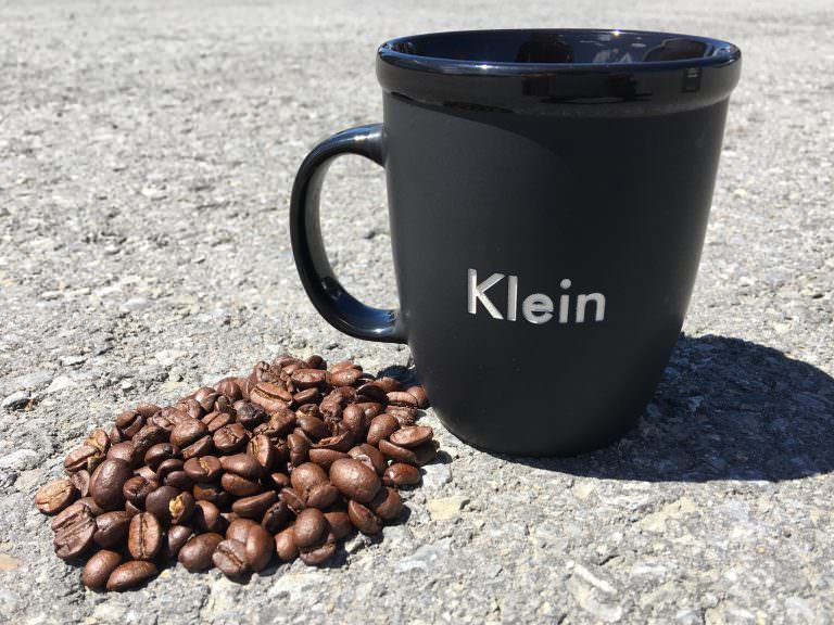 A pile of whole coffee beans next to a Klein 12 oz Ceramic Bistro Mug on asphalt.